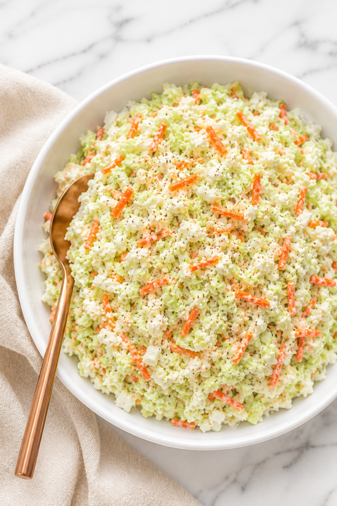 Creamy copycat KFC coleslaw in a white serving bowl with visible shredded carrots and celery seed garnish, styled on a white marble countertop with a neutral linen cloth and copper spoon.