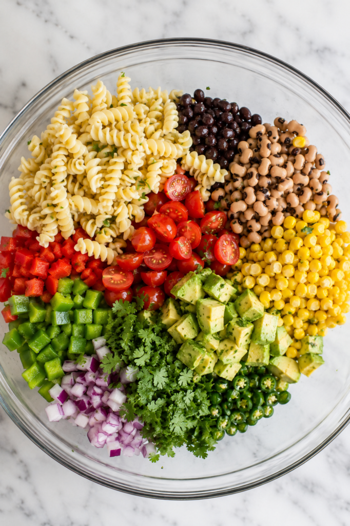 Cowboy caviar pasta salad ingredients layered in a large glass bowl before mixing on a white marble countertop