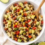Cowboy caviar pasta salad with rotini pasta, black beans, corn, avocado, tomatoes, and peppers in a white bowl on a marble countertop with a wooden serving spoon