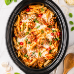 Crockpot meatball pasta bake with penne pasta, marinara sauce, mozzarella cheese, and basil in a black slow cooker on a white marble countertop