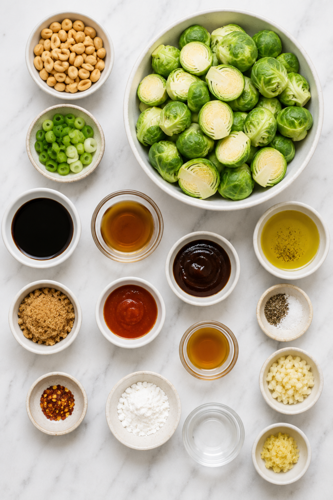 Flat lay of ingredients for kung pao Brussels sprouts including Brussels sprouts, soy sauce, peanuts, garlic, ginger, green onions, and seasonings on a marble countertop
