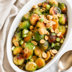 Kung pao Brussels sprouts in a white serving bowl topped with peanuts and green onions on a marble counter with a linen towel and serving spoon
