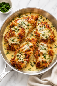 Crispy parmesan crusted chicken in creamy sauce served in a stainless steel skillet on a white marble countertop