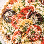 Unbaked old-fashioned tomato pie with heirloom tomatoes, sweet onions, fresh herbs, Gruyère, and Parmesan in a flaky homemade crust on a marble countertop.