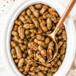 Southern crockpot boiled peanuts in a white slow cooker with a gold spoon scooping peanuts on a marble countertop with parsley and red pepper flakes