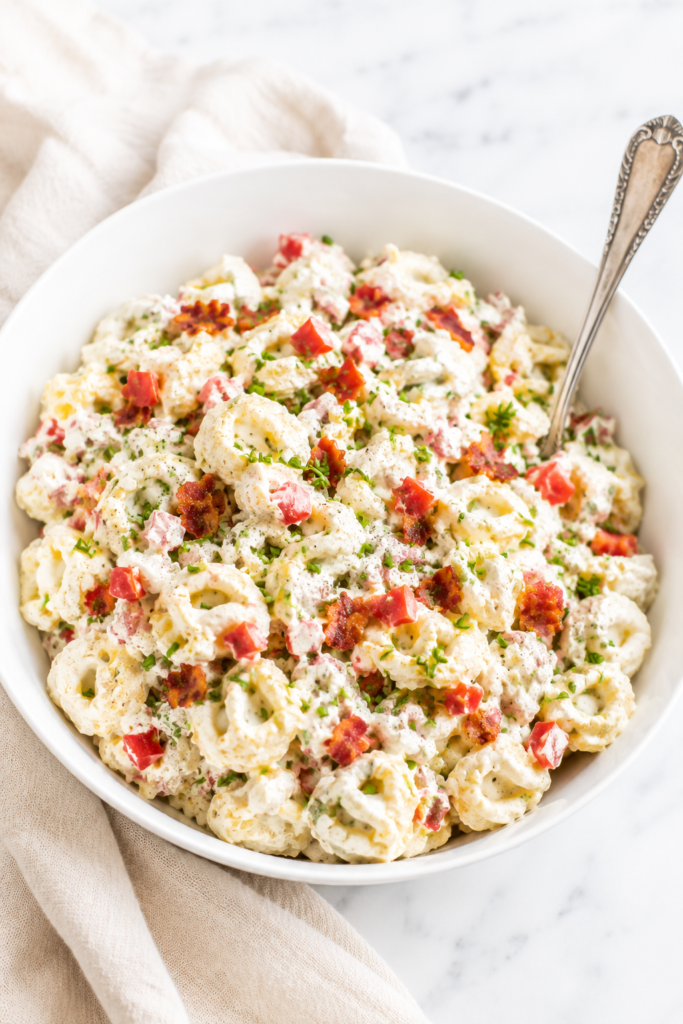 Creamy garlic BLT tortellini pasta salad in a white bowl topped with crispy bacon, diced tomatoes, chopped chives, and a rich garlic dressing on a marble countertop.