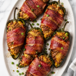 Bacon wrapped poblano poppers stuffed with creamy ground beef and cheese filling on a white oval platter over a white marble countertop.