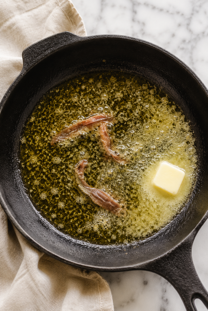 Anchovies melting into butter and olive oil in a cast iron skillet while preparing traditional bagna cauda sauce on a white marble countertop.