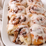 Soft and gooey Amish cinnamon rolls made with mashed potatoes and topped with creamy vanilla frosting on a white platter.