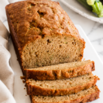 A sliced loaf of moist classic zucchini bread on a white marble countertop with a neutral linen towel and fresh zucchini.