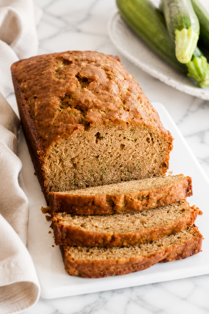 A sliced loaf of moist classic zucchini bread on a white marble countertop with a neutral linen towel and fresh zucchini.