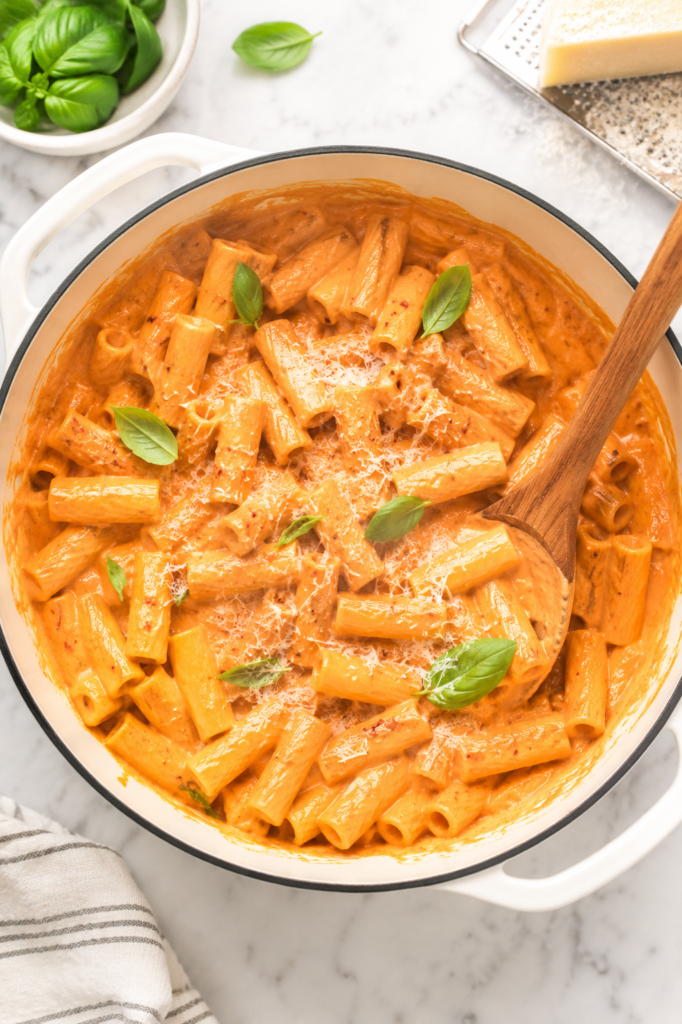 Creamy spicy rigatoni vodka pasta in a white enamel pot with Parmesan cheese and fresh basil on a marble countertop.