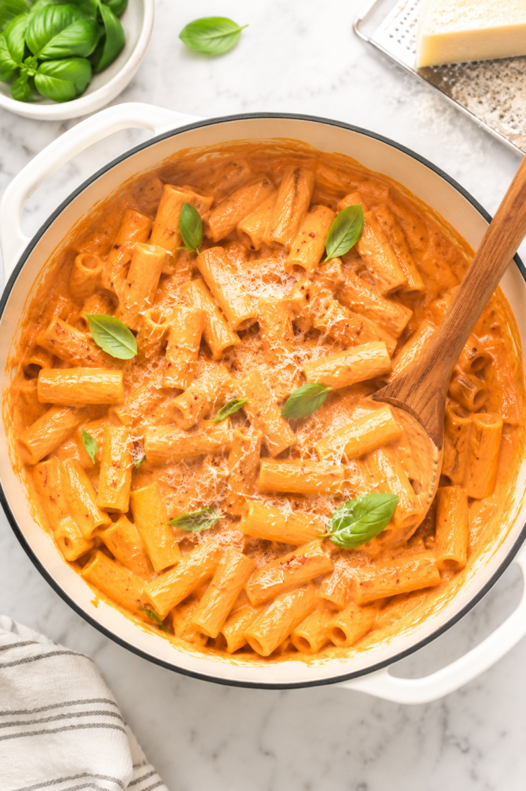 Creamy spicy rigatoni vodka pasta in a white enamel pot with Parmesan cheese and fresh basil on a marble countertop.