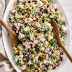 Creamy chicken bacon ranch broccoli salad served on a white oval platter with wooden salad tossers on a marble countertop.