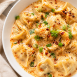 Chicken wontons in a creamy garlic chili crisp sauce served in a white bowl on a marble countertop with a neutral linen cloth.