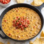 Creamy Chili's copycat skillet queso made with seasoned ground beef and melted cheese in a cast iron skillet on a white marble countertop with tortilla chips scattered around.