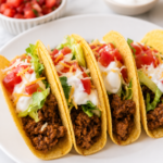 Four crunchy beef tacos topped with lettuce, tomatoes, shredded cheese, and sour cream on a white plate over a white marble countertop.