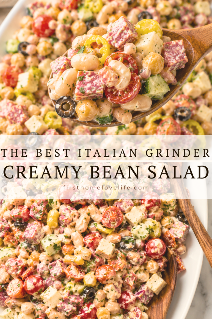 Wooden spoon lifting a scoop of creamy Italian Grinder Bean Salad with cannellini beans, chickpeas, salami, pepperoni, provolone cheese, cherry tomatoes, pepperoncini, olives, and cucumber from a white serving platter.