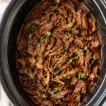 Crockpot beef barbacoa made with tender shredded chuck roast slow cooked in a smoky chipotle sauce inside a slow cooker on a white marble countertop.