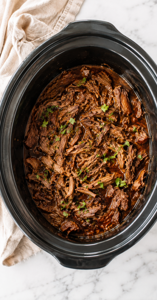 Crockpot beef barbacoa made with tender shredded chuck roast slow cooked in a smoky chipotle sauce inside a slow cooker on a white marble countertop.