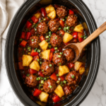 Bird's-eye view of Crockpot Hawaiian meatballs with pineapple chunks and sweet tangy sauce in an oval slow cooker on a white marble countertop.
