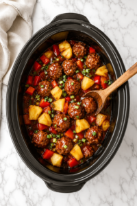 Bird's-eye view of Crockpot Hawaiian meatballs with pineapple chunks and sweet tangy sauce in an oval slow cooker on a white marble countertop.