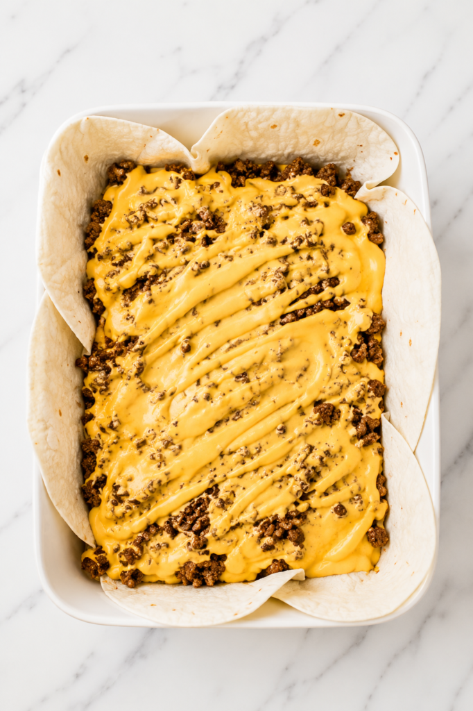 Overhead view of Crunchwrap Supreme Casserole being assembled in a white baking dish with flour tortillas, seasoned taco beef, and nacho cheese sauce on a marble countertop.