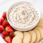 Creamy homemade Dunkaroo dip topped with colorful rainbow sprinkles in a white bowl, served with soft sugar cookies and fresh strawberries on a white marble countertop.