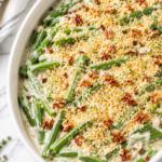 Creamy green bean goat cheese gratin topped with Parmesan breadcrumbs and chopped pecans in a white baking dish on a marble countertop.