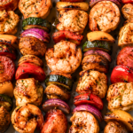 Grilled jambalaya kabobs with Cajun-seasoned chicken, shrimp, andouille sausage, bell peppers, onions, zucchini, and mushrooms on a white marble countertop.