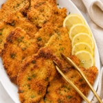 Golden crispy Italian breaded chicken cutlets arranged on a white platter with sliced lemons and gold tongs on a marble countertop.