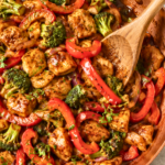 Jerk chicken stir fry with broccoli, red bell peppers, and onions cooked in a copper pan on a white marble countertop.