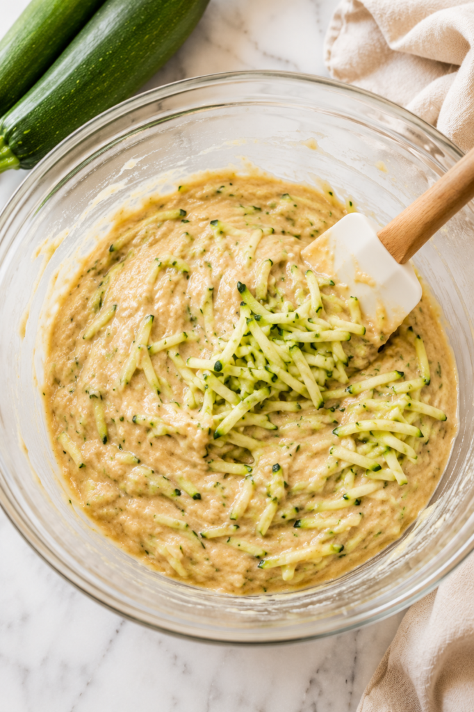 Fresh shredded zucchini being folded into homemade zucchini bread batter with a spatula in a large glass mixing bowl on a white marble countertop.