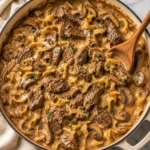 Creamy one pan steak Diane stroganoff with tender beef, mushrooms, and egg noodles in a white enamel skillet on a marble countertop with a wooden spoon.