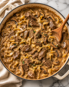 Creamy one pan steak Diane stroganoff with tender beef, mushrooms, and egg noodles in a white enamel skillet on a marble countertop with a wooden spoon.