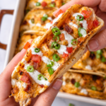 Hand holding a cheesy oven baked crack chicken taco topped with ranch dressing, bacon, diced tomatoes, and green onions with crispy baked tacos in the background.
