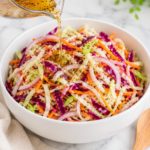 Pickling liquid being poured over a colorful cabbage slaw in a white bowl on a marble countertop with a wooden spoon and neutral linen cloth.