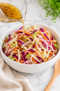 Pickling liquid being poured over a colorful cabbage slaw in a white bowl on a marble countertop with a wooden spoon and neutral linen cloth.