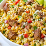 White bowl filled with steak and Mexican street corn pasta salad featuring grilled steak, roasted corn, cotija cheese, cilantro, lime wedges, and creamy chili lime dressing on a white marble countertop.