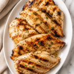 Juicy grilled chicken breasts on a white oval platter with grill marks, served on a marble countertop with a neutral linen cloth.
