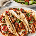 Crispy tortilla pizza tacos stuffed with seasoned beef, pizza sauce, melted mozzarella cheese, and pepperoni on a white platter with a fresh Italian salad topped with Parmesan on a marble countertop.