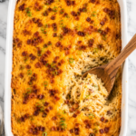 Creamy Waffle House loaded hashbrown casserole with melted cheddar cheese, crispy bacon, and green onions in a white baking dish with a wooden spoon serving a scoop from the casserole.