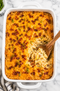 Creamy Waffle House loaded hashbrown casserole with melted cheddar cheese, crispy bacon, and green onions in a white baking dish with a wooden spoon serving a scoop from the casserole.