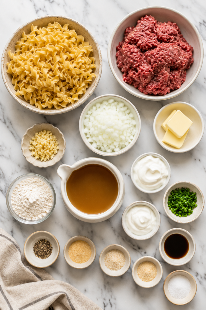 Flat lay of ingredients for Amish Style Beef and Noodles including ground beef, Amish egg noodles, beef broth, onion, garlic, butter, sour cream, heavy cream, flour, Worcestershire sauce, chopped chives, and seasonings on a white marble countertop.