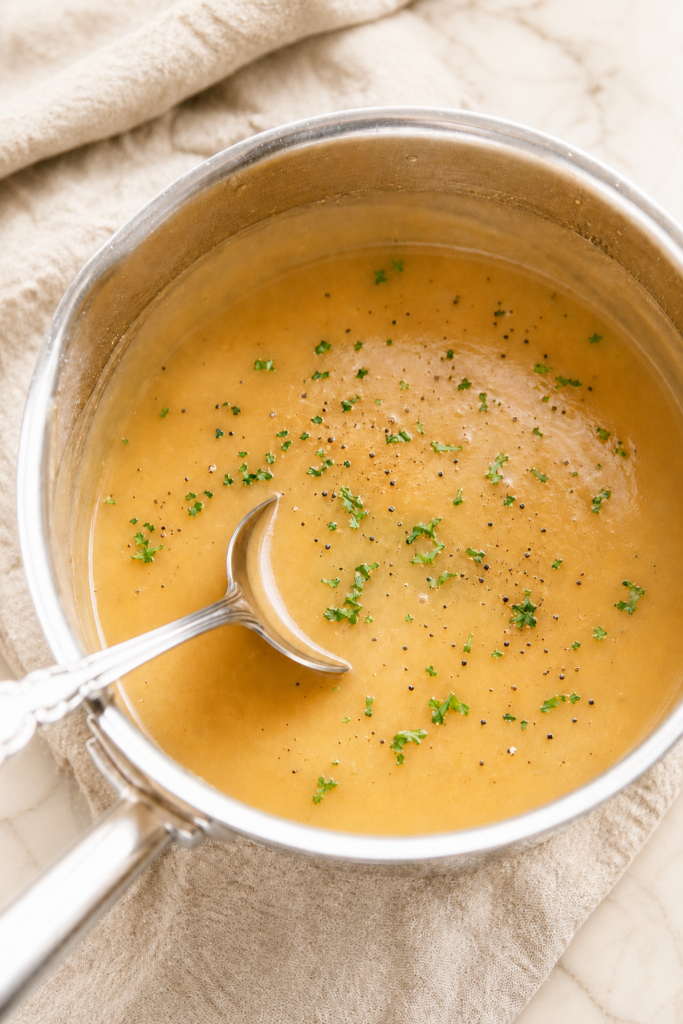 Homemade turkey gravy from scratch with pan drippings, parsley, and cracked black pepper in a stainless steel saucepan with a silver ladle.