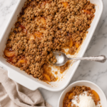 Bird's-eye view of homemade brown butter peach crisp in a white rectangular baking dish with a golden oat crumble topping. A serving is scooped into a small white bowl with melting vanilla ice cream on a marble countertop and a neutral kitchen towel.
