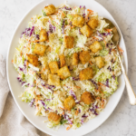 Caesar salad coleslaw made with green and red cabbage, carrots, Parmesan cheese, creamy Caesar dressing, and crunchy croutons on a white platter.