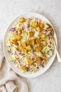 Caesar salad coleslaw made with green and red cabbage, carrots, Parmesan cheese, creamy Caesar dressing, and crunchy croutons on a white platter.