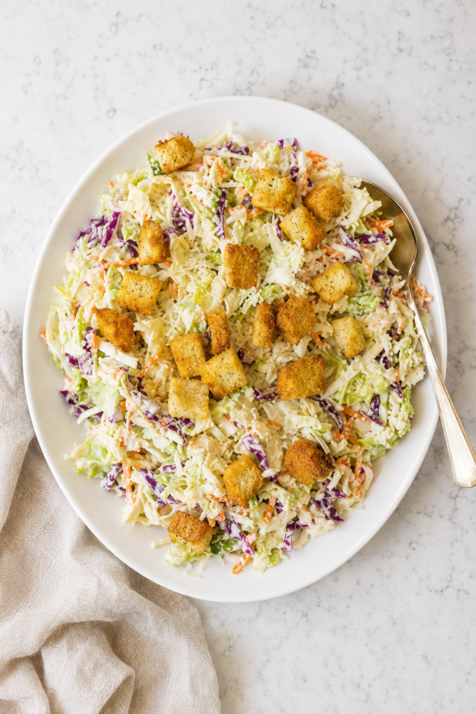 Caesar salad coleslaw made with green and red cabbage, carrots, Parmesan cheese, creamy Caesar dressing, and crunchy croutons on a white platter.