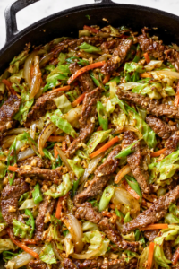 Chinese beef and cabbage stir fry with sliced steak, cabbage, carrots, onions, and savory garlic ginger sauce in a cast-iron skillet.